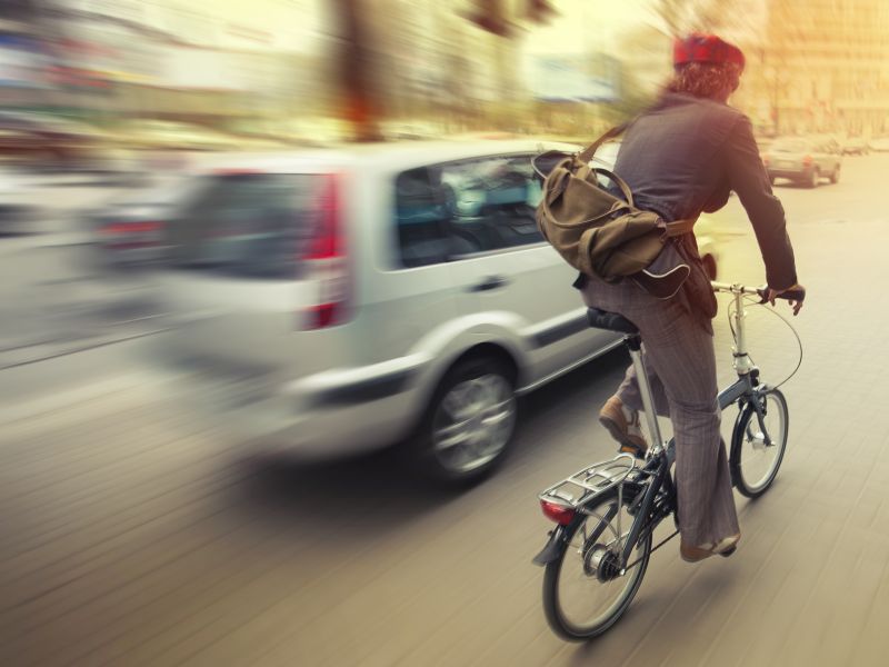 Ein Radfahrer fährt rechts auf einem Klapprad auf der Straße und wird gerade von einem weißen Auto links überholt.