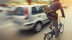 Ein Radfahrer fährt rechts auf einem Klapprad auf der Straße und wird gerade von einem weißen Auto links überholt.