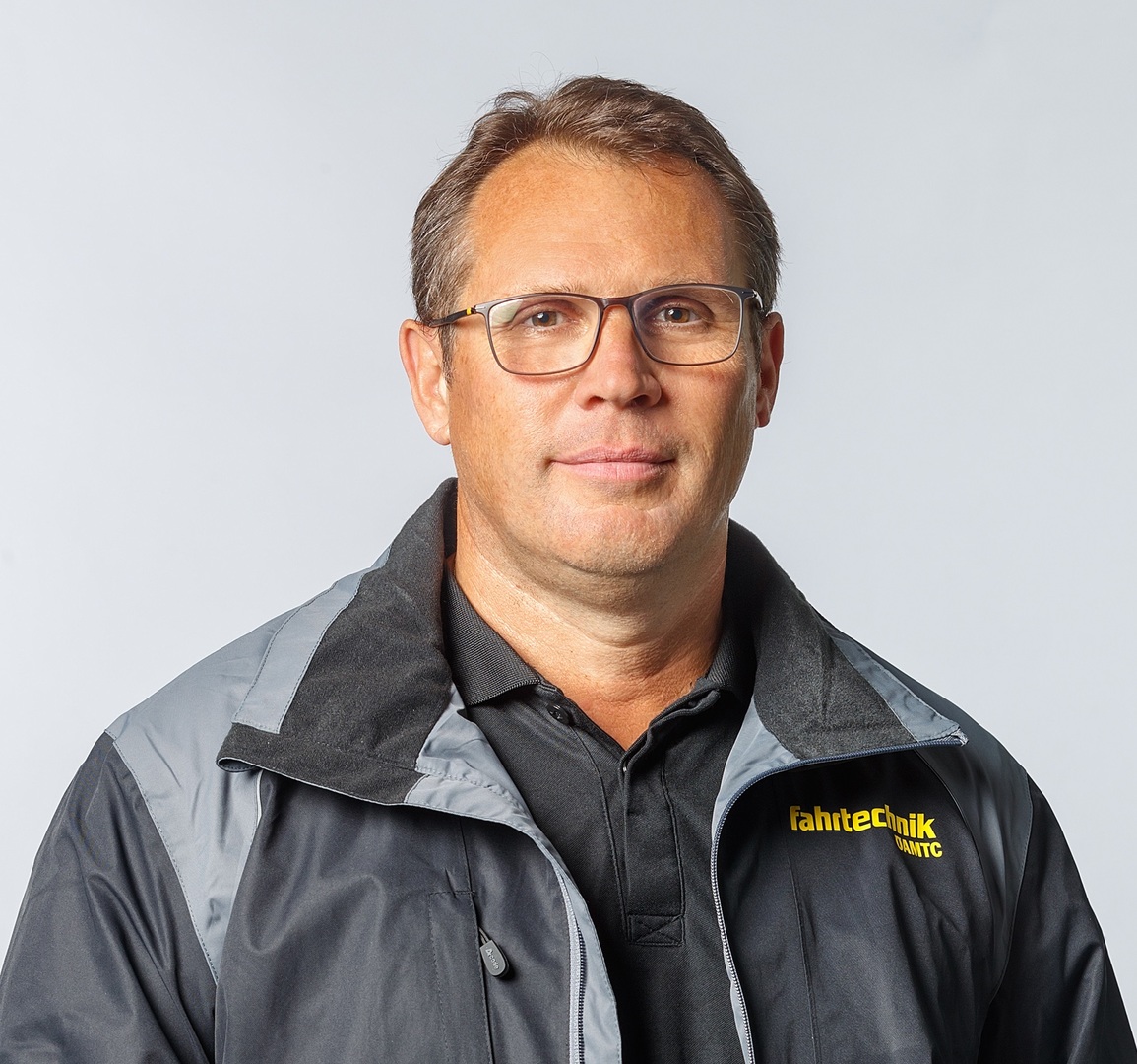 Das Bild ist ein Portrait von Roland Frisch, der frontal in die Kamera schaut und eine schwarze Jacke mit gelber ÖAMTC Fahrtechnik-Aufschrift trägt.
