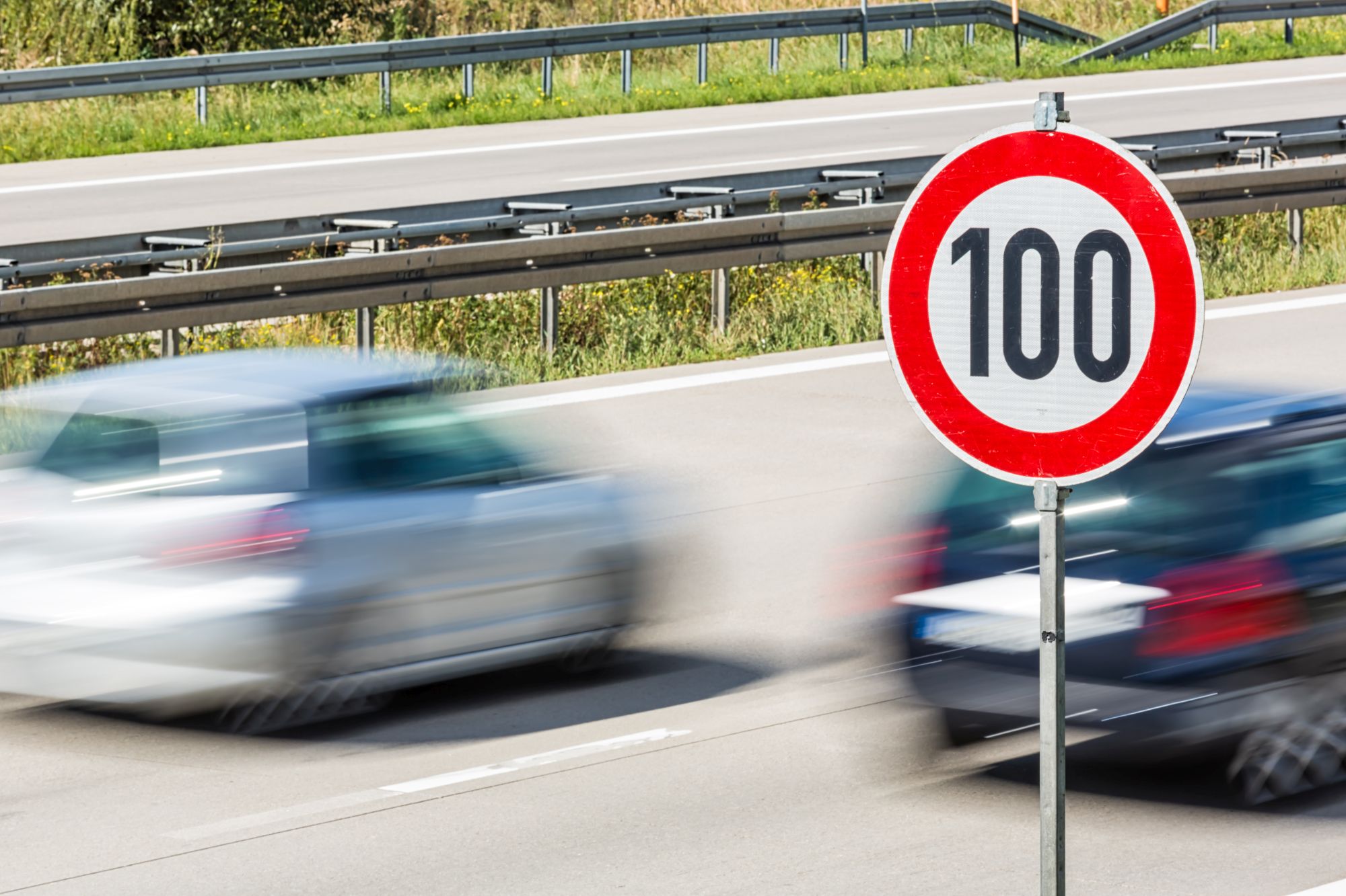 Zwei Autos mit hoher Geschwindigkeit auf einer Autobahn. Im Vordergrund ein Verkehrsschild, das eine 100 km/h Beschränkung anzeigt.