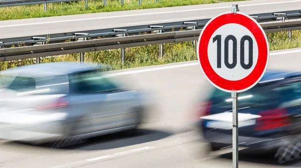 Zwei Autos mit hoher Geschwindigkeit auf einer Autobahn. Im Vordergrund ein Verkehrsschild, das eine 100 km/h Beschränkung anzeigt.
