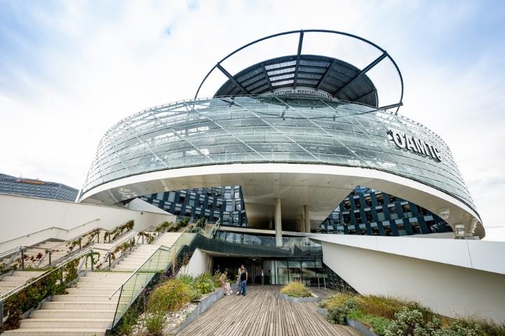 Das Mobilitätszentrum von aussen. Das Gebäude ist rund und die Front aus Glas. im Vordergrund sieht man eine teilweise befpflanzte Terrasse mit einem Holzboden und einen Stiegenaufgang.