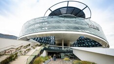 Das Mobilitätszentrum von aussen. Das Gebäude ist rund und die Front aus Glas. im Vordergrund sieht man eine teilweise befpflanzte Terrasse mit einem Holzboden und einen Stiegenaufgang.