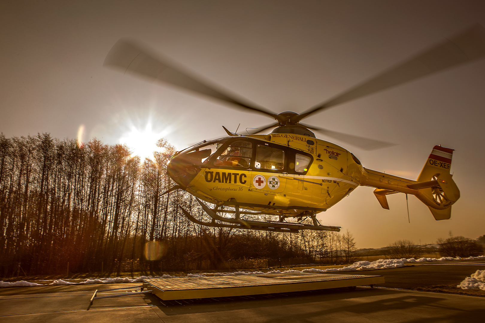 Christophorus-Notarzthubschrauber, der gerade abhebt oder landet. Die Sonne steht tief am Himmel und erzeugt eine warm-goldene Beleuchtung, während die Rotorblätter des Hubschraubers sich drehen.