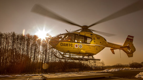 Christophorus-Notarzthubschrauber, der gerade abhebt oder landet. Die Sonne steht tief am Himmel und erzeugt eine warm-goldene Beleuchtung, während die Rotorblätter des Hubschraubers sich drehen.