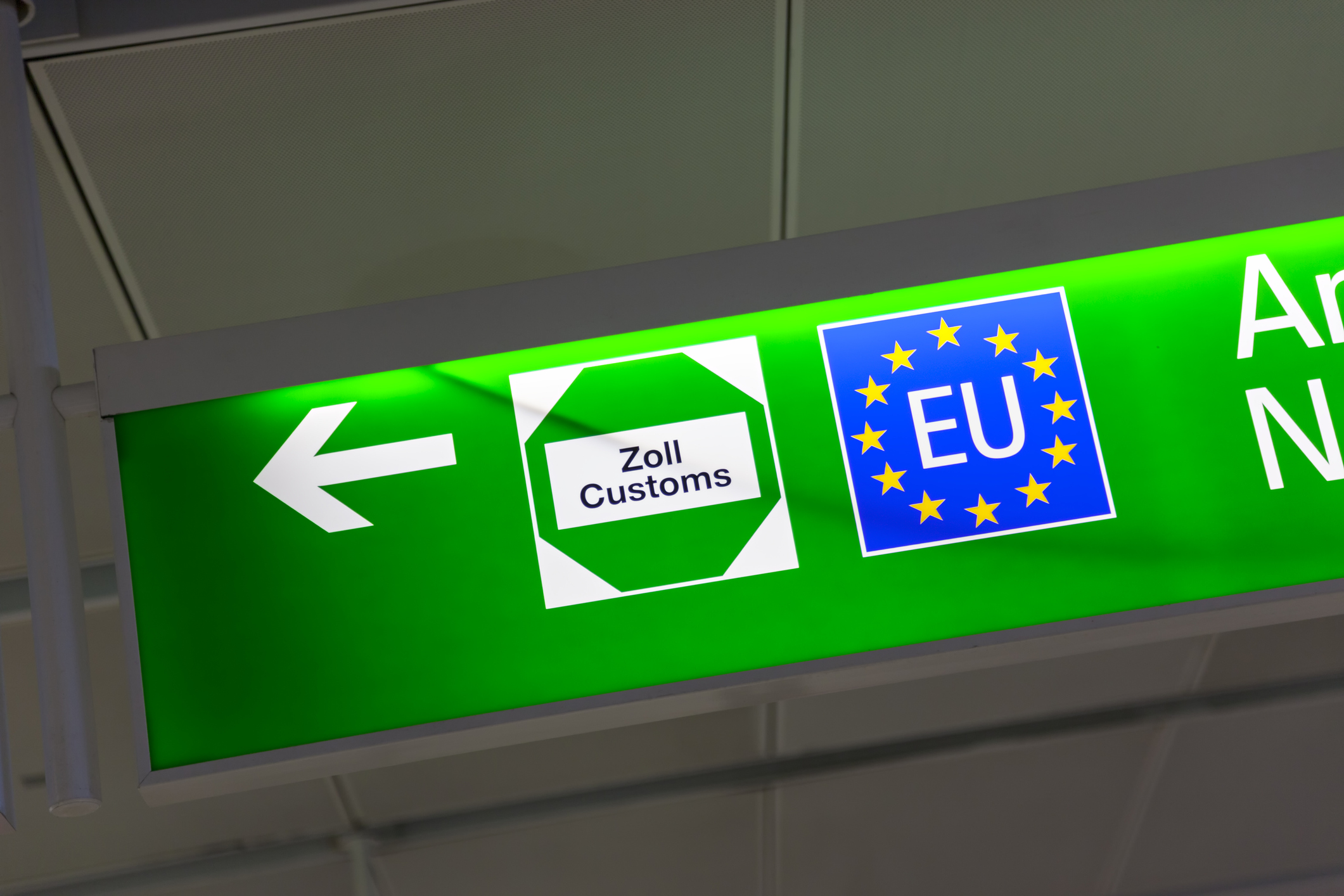 Ein grünes Schild mit einem weißen Pfeil nach links. Auf dem Schild sind zwei Symbole abgebildet: links ein weißes Sechseck mit der Aufschrift "Zoll Customs"  und rechts ein blaues Quadrat mit gelben Sternen, dass die Flagge der EU darstellt.