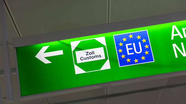 Ein grünes Schild mit einem weißen Pfeil nach links. Auf dem Schild sind zwei Symbole abgebildet: links ein weißes Sechseck mit der Aufschrift "Zoll Customs" und rechts ein blaues Quadrat mit gelben Sternen, dass die Flagge der EU darstellt.