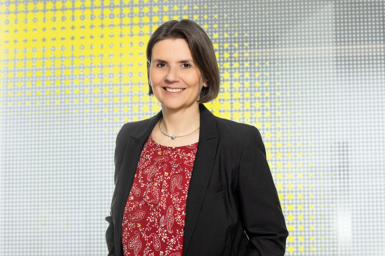 Porträtfoto von einer Frau mit kinnlangen braunen Haaren, die ein rotes Shirt mit weißem Muster und einen schwarzen Balzer trägt und vor einer gelben-weißen Glaswand steht.