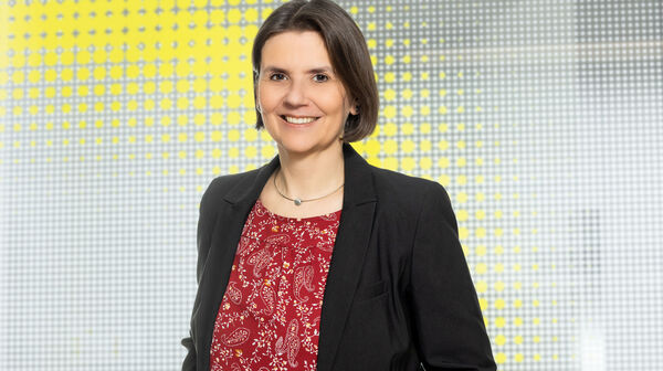 Porträtfoto von einer Frau mit kinnlangen braunen Haaren, die ein rotes Shirt mit weißem Muster und einen schwarzen Balzer trägt und vor einer gelben-weißen Glaswand steht.