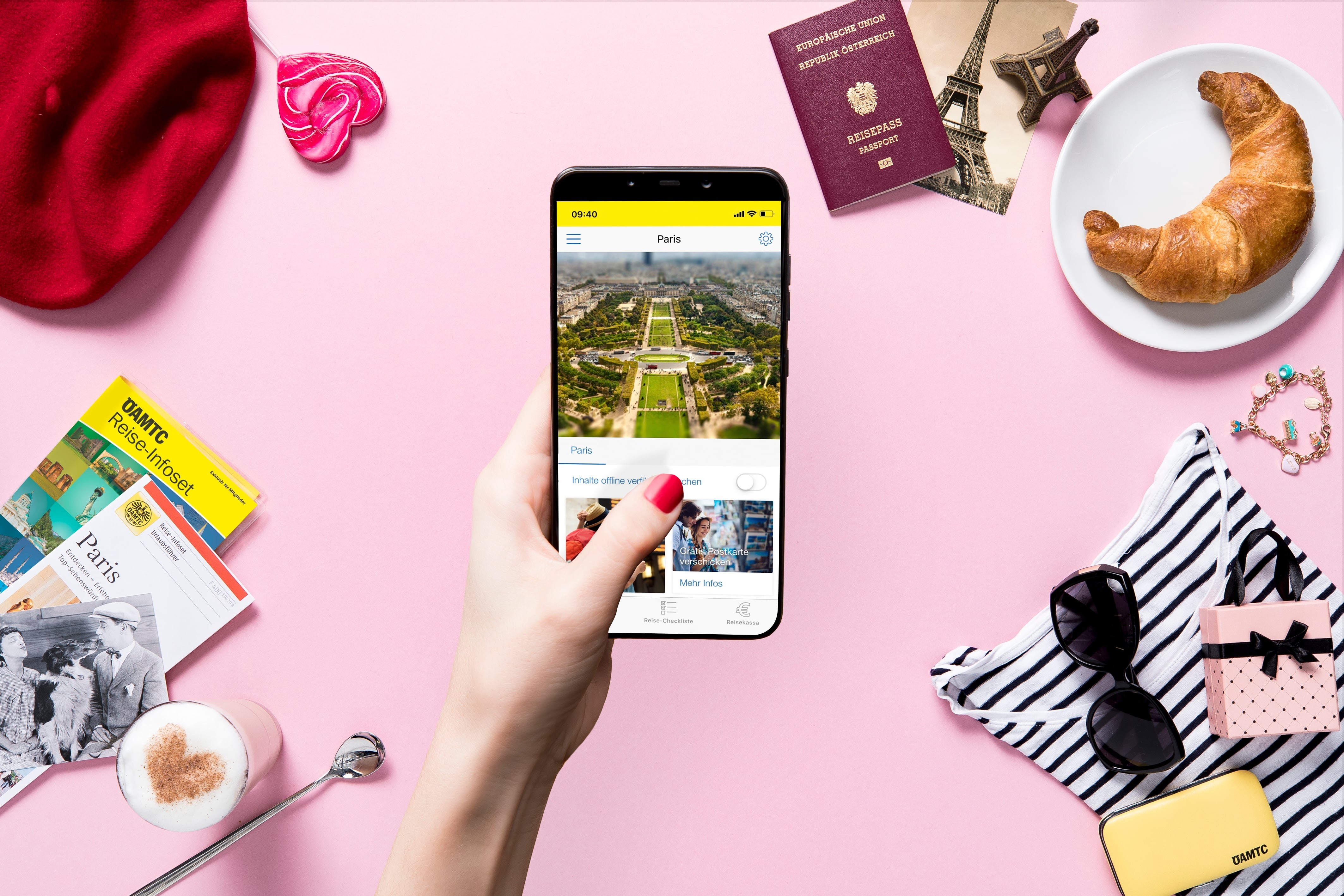 Eine Hand hält ein Smartphone mit einem Bild von Paris. Im pinkfärbigen Hintergrund liegen ein Reiseführer, ein Reisepass, ein Croissant, eine Sonnenbrille, ein Herz-Macchiato, eine rote Baskenmütze und weitere Reiseutensilien.
