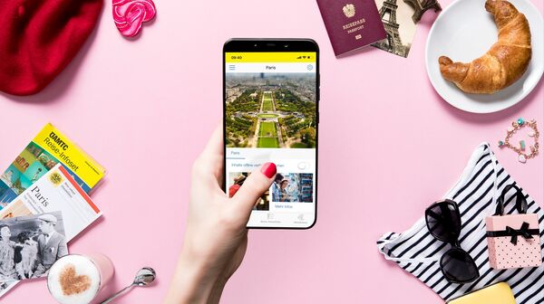 Eine Hand hält ein Smartphone mit einem Bild von Paris. Im pinkfärbigen Hintergrund liegen ein Reiseführer, ein Reisepass, ein Croissant, eine Sonnenbrille, ein Herz-Macchiato, eine rote Baskenmütze und weitere Reiseutensilien.