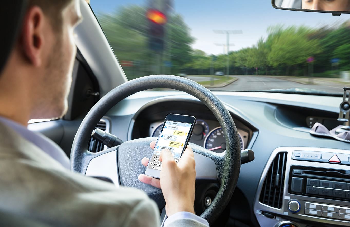 Ein Mann sitzt in einem Auto, in seiner rechten Hand hält er ein Smartphone. Sein Blick ist auf das Handy gerichtet, während das Lenkrad mit der linken Hand gehalten wird. Im Hintergrund sind das Armaturenbrett und Teile der Windschutzscheibe zu sehen. Die Szene vermittelt den Eindruck, dass die Person während der Fahrt durch das Handy abgelenkt ist.