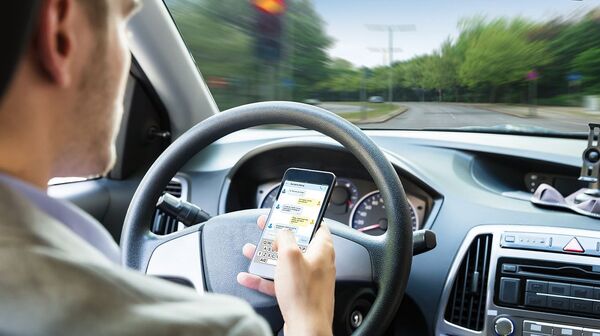Ein Mann sitzt in einem Auto, in seiner rechten Hand hält er ein Smartphone. Sein Blick ist auf das Handy gerichtet, während das Lenkrad mit der linken Hand gehalten wird. Im Hintergrund sind das Armaturenbrett und Teile der Windschutzscheibe zu sehen. Die Szene vermittelt den Eindruck, dass die Person während der Fahrt durch das Handy abgelenkt ist.