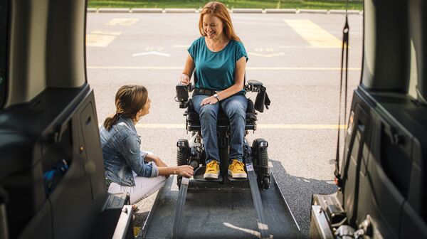 Eine Frau sitzt in einem elektrischen Rollstuhl und fährt eine Rampe in ein größeres Auto hoch. Eine weitere Frau ist neben der Rampe behilflich.