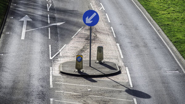 Eine Straße an der Linksverkehr herrscht. Eine Straßenschild auf dem ein Pfeil abgebildet ist, der nach links unten zeigt.