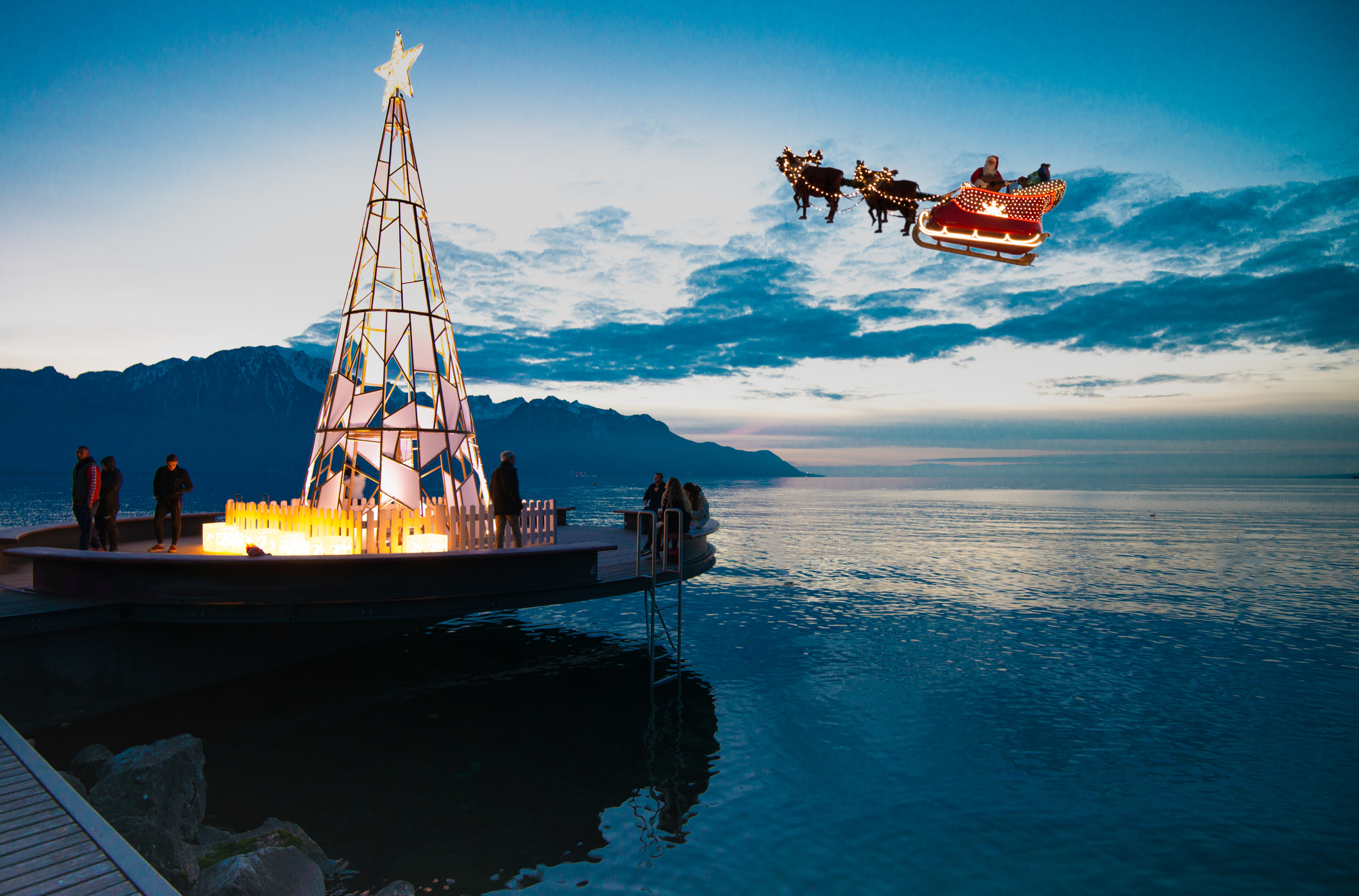 Ein beleuchteter Weihnachtsbaum auf einem Steg am Meer, mit einem fliegenden Weihnachtsmannschlitten im Hintergrund.