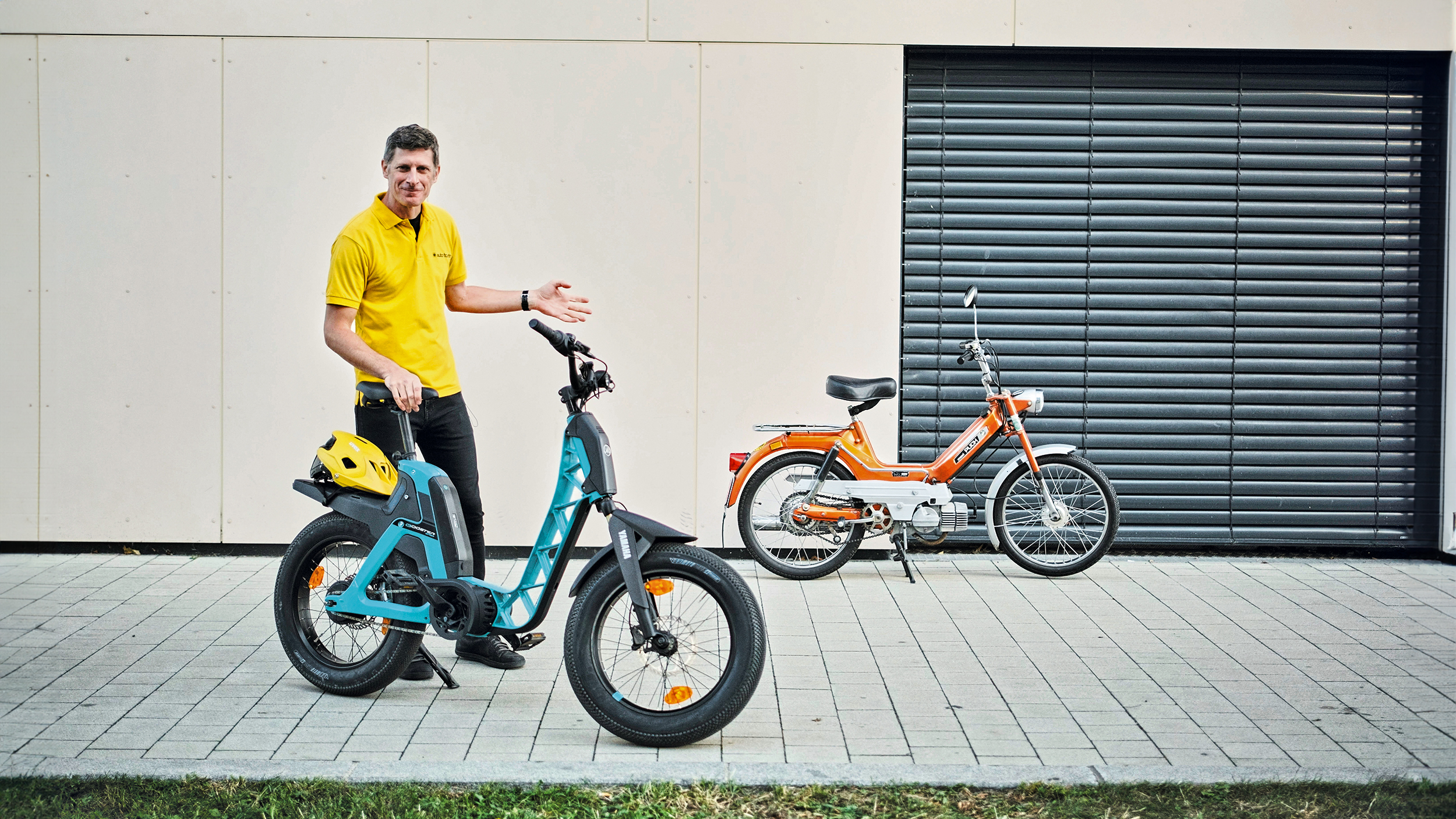 auto touring-Redakteur Alex Fischer steht zwischen zwei Zweirädern. Links befindet sich das moderne, blaue Elektrofahrrad, die Yamaha Booser easy, mit einem gelben Helm auf dem Gepäckträger. Rechts steht das klassische, orangefarbene Moped – die Puch Maxi.