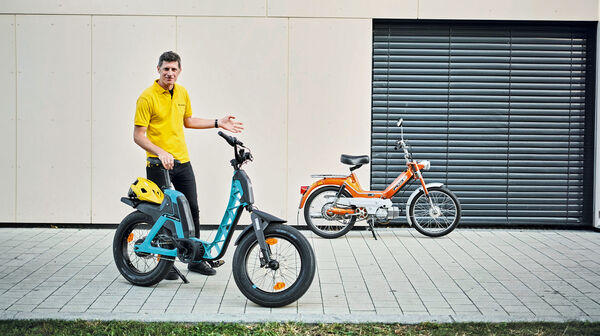 auto touring-Redakteur Alex Fischer steht zwischen zwei Zweirädern. Links befindet sich das moderne, blaue Elektrofahrrad, die Yamaha Booser easy, mit einem gelben Helm auf dem Gepäckträger. Rechts steht das klassische, orangefarbene Moped – die Puch Maxi.