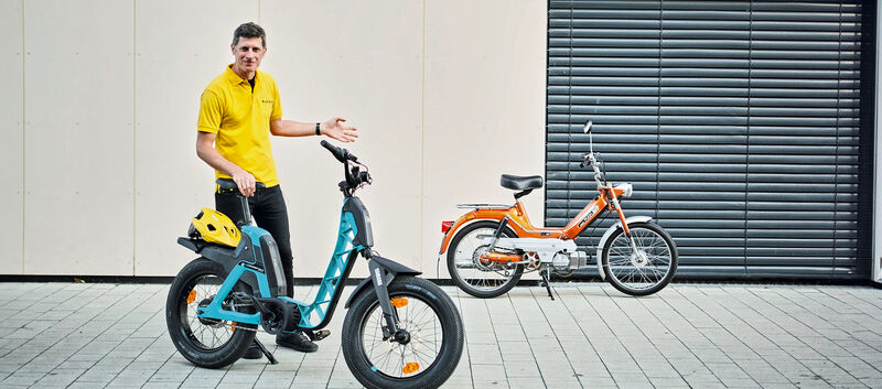 auto touring-Redakteur Alex Fischer steht zwischen zwei Zweirädern. Links befindet sich das moderne, blaue Elektrofahrrad, die Yamaha Booser easy, mit einem gelben Helm auf dem Gepäckträger. Rechts steht das klassische, orangefarbene Moped – die Puch Maxi.