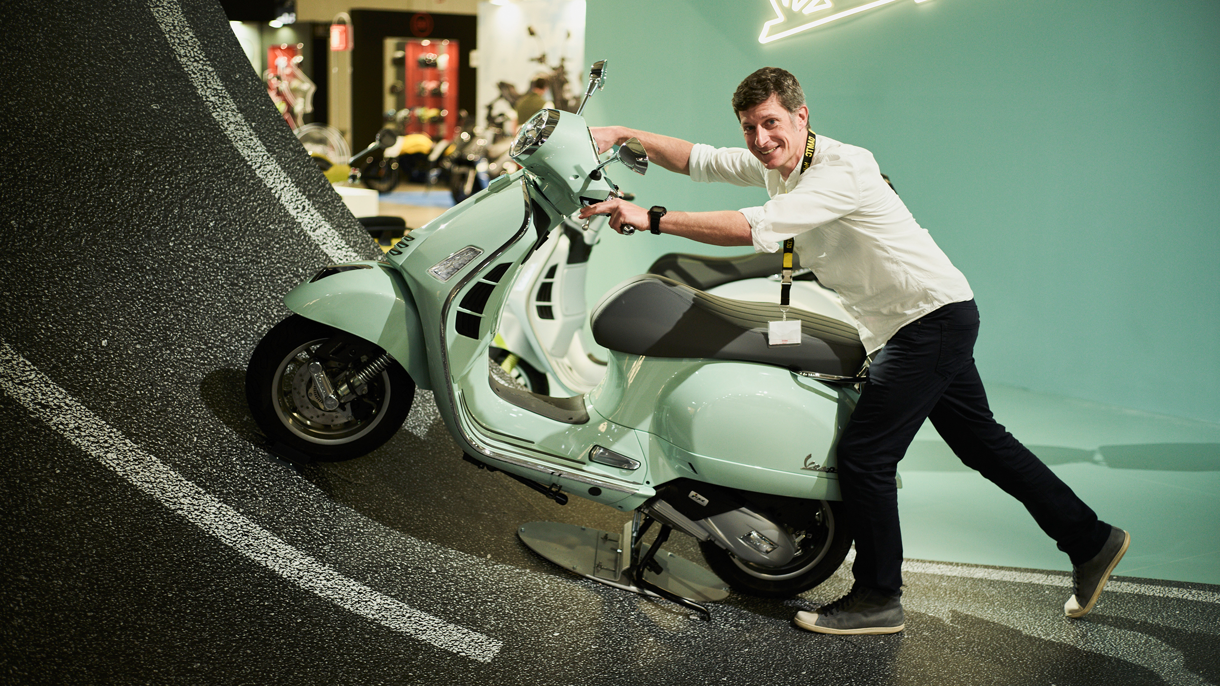 An einem Messestand steht auf einer steilen Fahrbahn eine mintgrüne Vespa. Ein Mann lacht und versucht sie die Fahrbahn hochzuschieben.