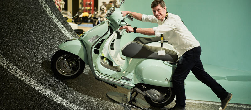 An einem Messestand steht auf einer steilen Fahrbahn eine mintgrüne Vespa. Ein Mann lacht und versucht sie die Fahrbahn hochzuschieben.