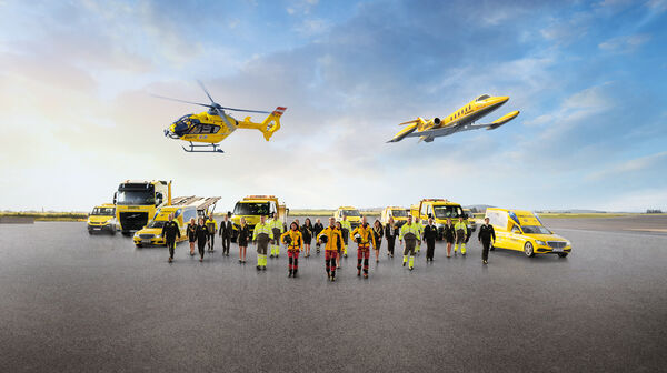 Eine Flotte von gelben Rettungsfahrzeugen und Luftfahrzeugen steht auf einem Flugfeld. Zu sehen sind mehrere Krankenwagen, ein Geländewagen, ein LKW, ein Rettungshubschrauber in der Luft sowie ein Ambulanzflugzeug. Davor stehen Einsatzkräfte in leuchtender Schutzkleidung.