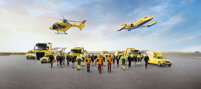 Eine Flotte von gelben Rettungsfahrzeugen und Luftfahrzeugen steht auf einem Flugfeld. Zu sehen sind mehrere Krankenwagen, ein Geländewagen, ein LKW, ein Rettungshubschrauber in der Luft sowie ein Ambulanzflugzeug. Davor stehen Einsatzkräfte in leuchtender Schutzkleidung.
