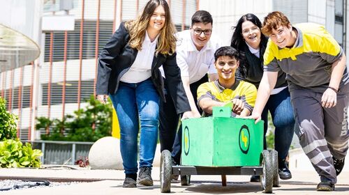 Vier Jugendliche schieben einen fünften Jugendlichen in einer Seifenkiste vor sich her auf die Kamera zu. Die Seifenkiste ist grün bemalt. Die Jugendlichen tragen ÖAMTC Dienstkleidung, sowohl für die Kfz-Technik als auch für den Schalterdienst. Im Hintergrund Pflanzen und Gebäude.