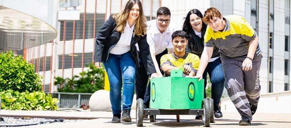 Vier Jugendliche schieben einen fünften Jugendlichen in einer Seifenkiste vor sich her auf die Kamera zu. Die Seifenkiste ist grün bemalt. Die Jugendlichen tragen ÖAMTC Dienstkleidung, sowohl für die Kfz-Technik als auch für den Schalterdienst. Im Hintergrund Pflanzen und Gebäude.
