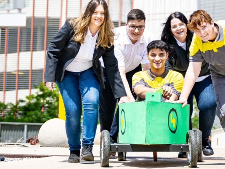 Vier Jugendliche schieben einen fünften Jugendlichen in einer Seifenkiste vor sich her auf die Kamera zu. Die Seifenkiste ist grün bemalt. Die Jugendlichen tragen ÖAMTC Dienstkleidung, sowohl für die Kfz-Technik als auch für den Schalterdienst. Im Hintergrund Pflanzen und Gebäude.
