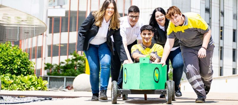 Vier Jugendliche schieben einen fünften Jugendlichen in einer Seifenkiste vor sich her auf die Kamera zu. Die Seifenkiste ist grün bemalt. Die Jugendlichen tragen ÖAMTC Dienstkleidung, sowohl für die Kfz-Technik als auch für den Schalterdienst. Im Hintergrund Pflanzen und Gebäude.