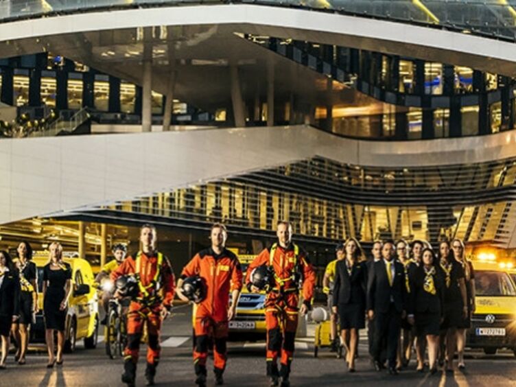 Die ÖAMTC Flotte vor dem Mobilitätszentrum. Viele Personen in Dienstkleidung: Kfz-Techniker:innen, Pilot:innen, Schalterdienstmitarbeiter:innen.