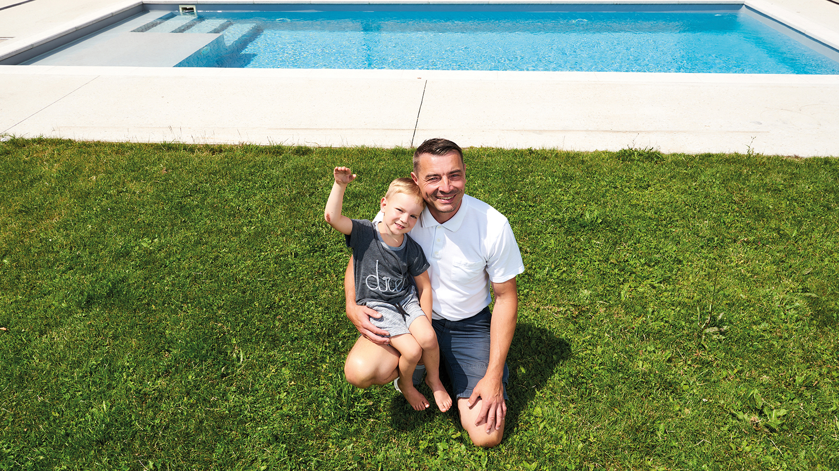 Emil kerngesund und winkend mit seinem Vater im Rasen vor dem Pool.
