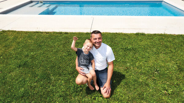 Emil kerngesund und winkend mit seinem Vater im Rasen vor dem Pool.