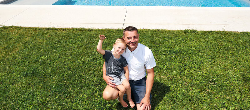 Emil kerngesund und winkend mit seinem Vater im Rasen vor dem Pool.