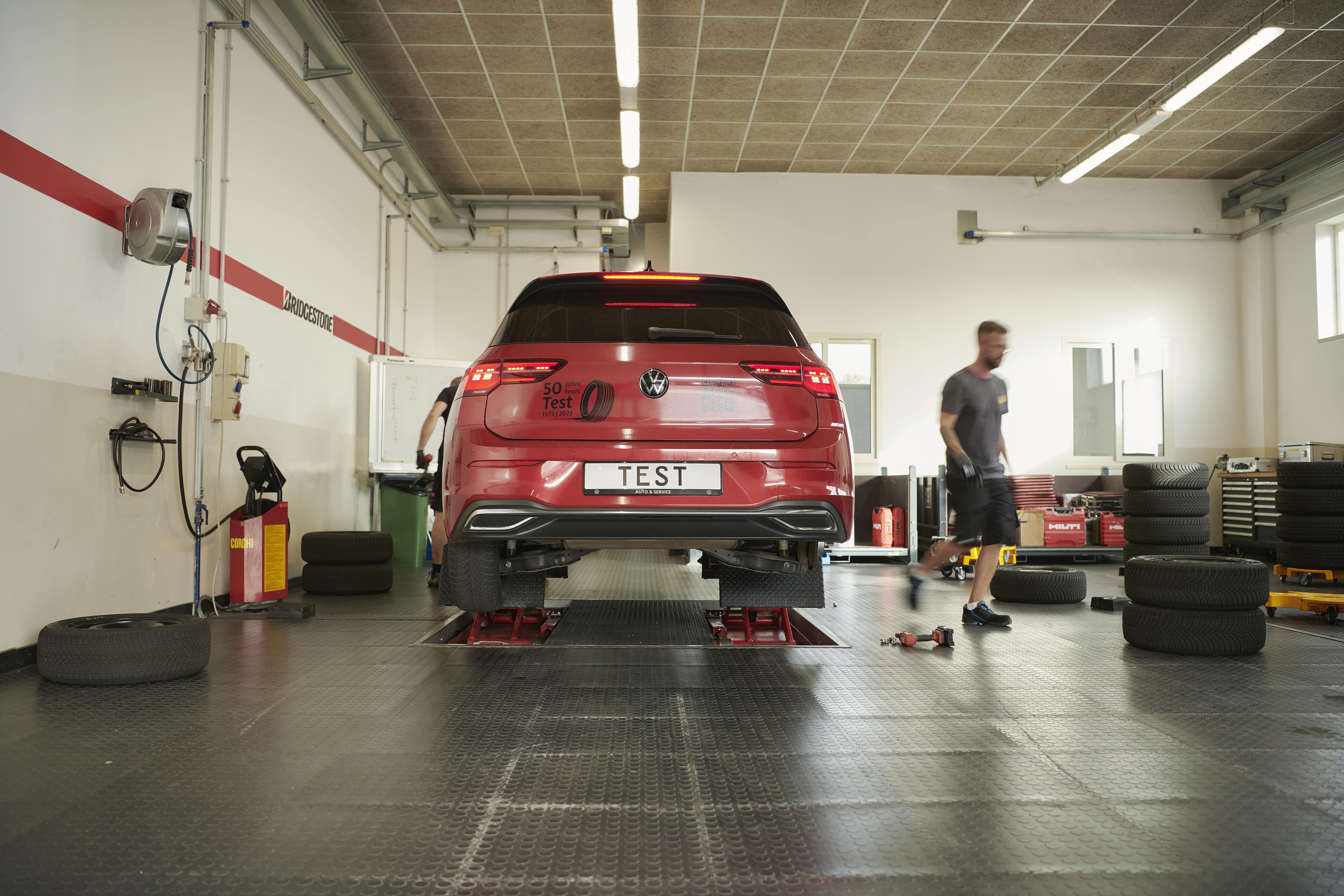 Sommerreifentest - Auto in Werkstatt ist auf der Hebebühne, rechts im Bild sieht man einzelne Reifen, Werkzeug und einen Mann.