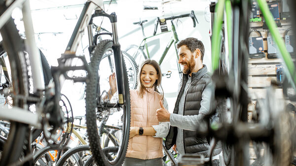 Ein junger Mann und eine junge Frau sehen sich in einem Fahrradgeschäft ein Fahrrad an. Beide lachen und der Mann gibt das Daumen-hoch-Zeichen.