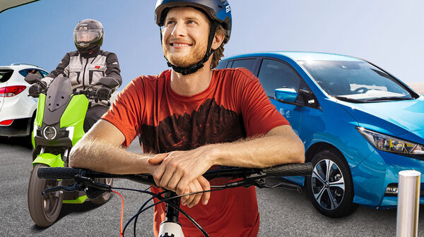 Ein Radfahrer mit Helm, ein E-Mopedfahrer mit Helm und ein blaues E-Auto beim Laden an einer Tankstelle. Im Hintergrund ist eine Wasserstoff Lademöglichkeit.