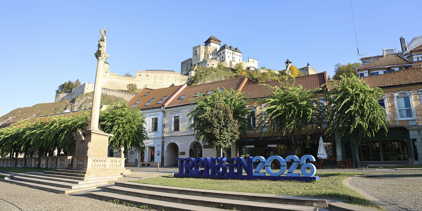 Der Hauptplatz mit einer Säule und einem blauen großen Schriftzug "Trencin 2026", dahinter auf einem Hügel eine Burg.
