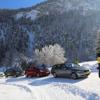 Im Bild sind vier Autos zu sehen, die auf einer Schneefahrbahn parken. Die Lenkerinnen und Lenker steigen aus und schauen zu einem Fahrtechnikinstruktor. Im Hintergrund sind Berge und Wälder zu sehen.