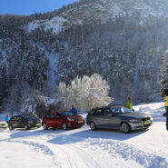 Im Bild sind vier Autos zu sehen, die auf einer Schneefahrbahn parken. Die Lenkerinnen und Lenker steigen aus und schauen zu einem Fahrtechnikinstruktor. Im Hintergrund sind Berge und Wälder zu sehen.