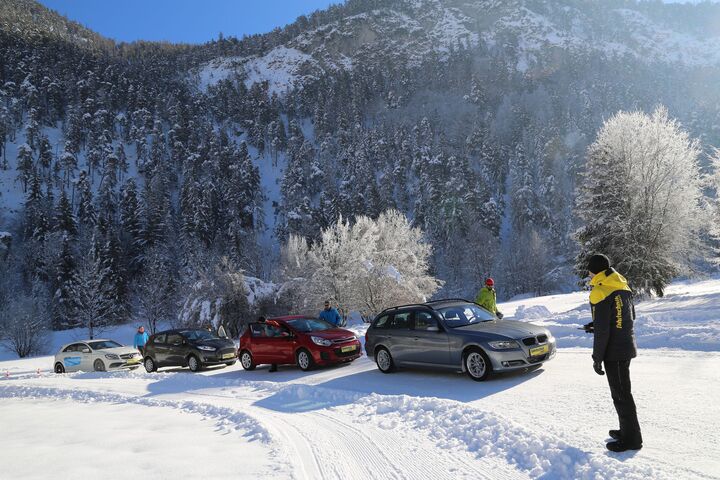 Im Bild sind vier Autos zu sehen, die auf einer Schneefahrbahn parken. Die Lenkerinnen und Lenker steigen aus und schauen zu einem Fahrtechnikinstruktor. Im Hintergrund sind Berge und Wälder zu sehen.