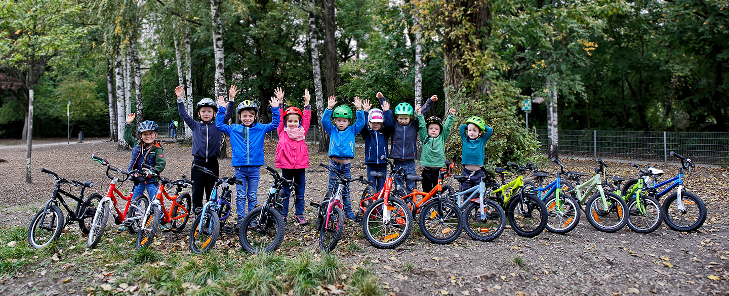 14 Kinderfahrräder sind auf einem Waldweg aufgestellt, dahinter einige Kinder mit Radhelm, die die Hände in die Höhe heben.