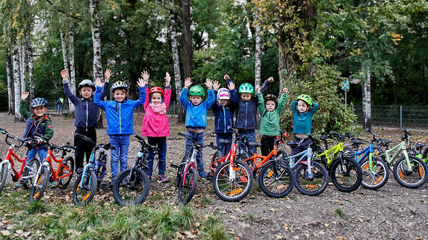 14 Kinderfahrräder sind auf einem Waldweg aufgestellt, dahinter einige Kinder mit Radhelm, die die Hände in die Höhe heben.
