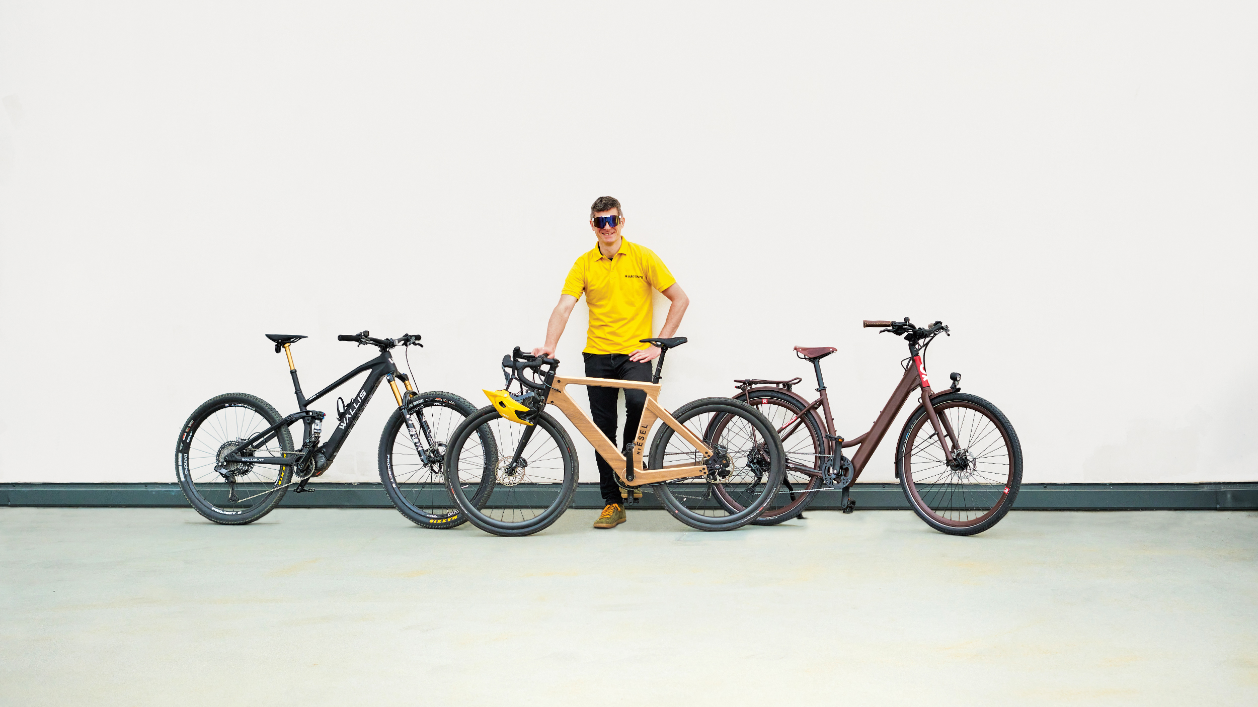 auto touring-Redakteur mit 3 E-Bikes unter 20 kg vor weißer Wand. Das Wallis Project S, das MyEsel E-Gravel und das Kraftstoff E-Razzo Wave.