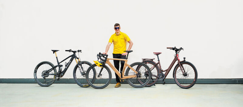 auto touring-Redakteur mit 3 E-Bikes unter 20 kg vor weißer Wand. Das Wallis Project S, das MyEsel E-Gravel und das Kraftstoff E-Razzo Wave.