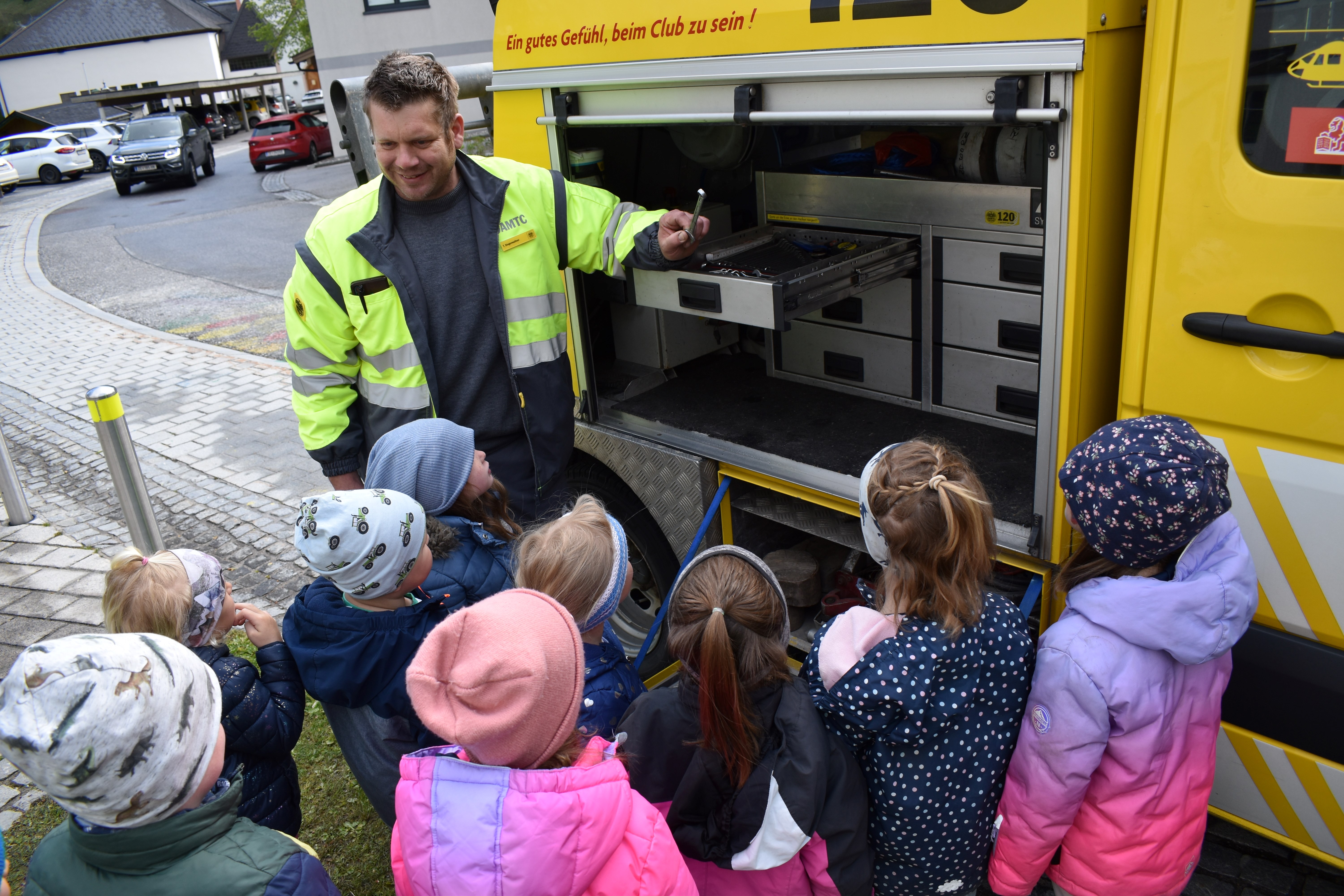 Pannenfahrer Thomas steht vor einer Gruppe an Kindergartenkinder und deutet auf den Inhalt seines Pannenfahrzeuges. 