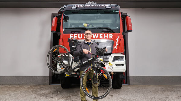 Feuerwehrmann Hannes Heiss steht vor einem Feuerwehrauto. Neben ihm sein Fahrrad.