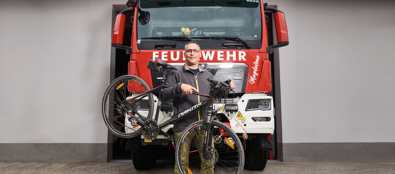 Feuerwehrmann Hannes Heiss steht vor einem Feuerwehrauto. Neben ihm sein Fahrrad.