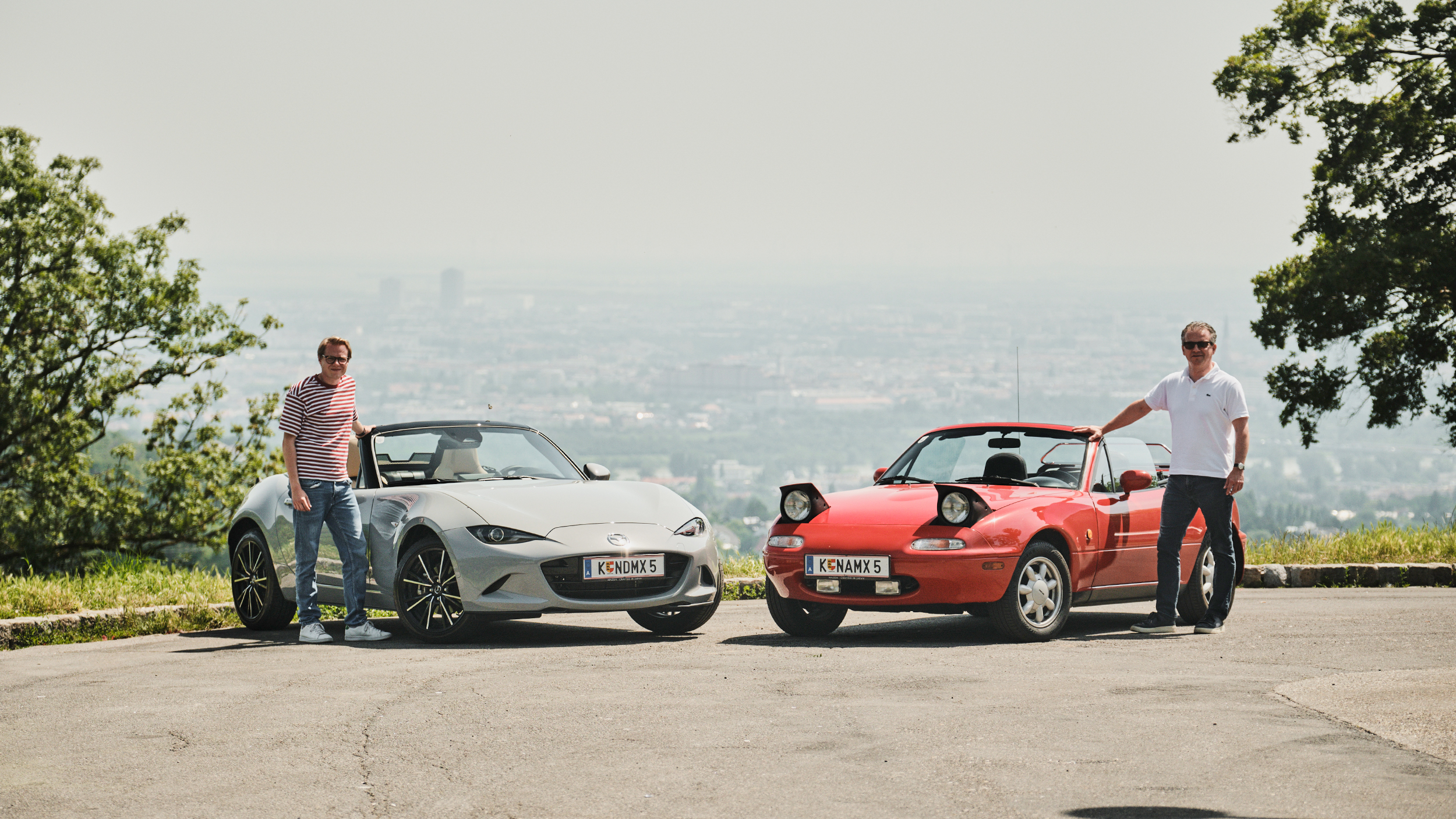 Zwei Personen stehen neben zwei Cabrios, der eine neben einem modernen, der andere neben einem alten. Im Hintergrund ist eine Stadt zu sehen.